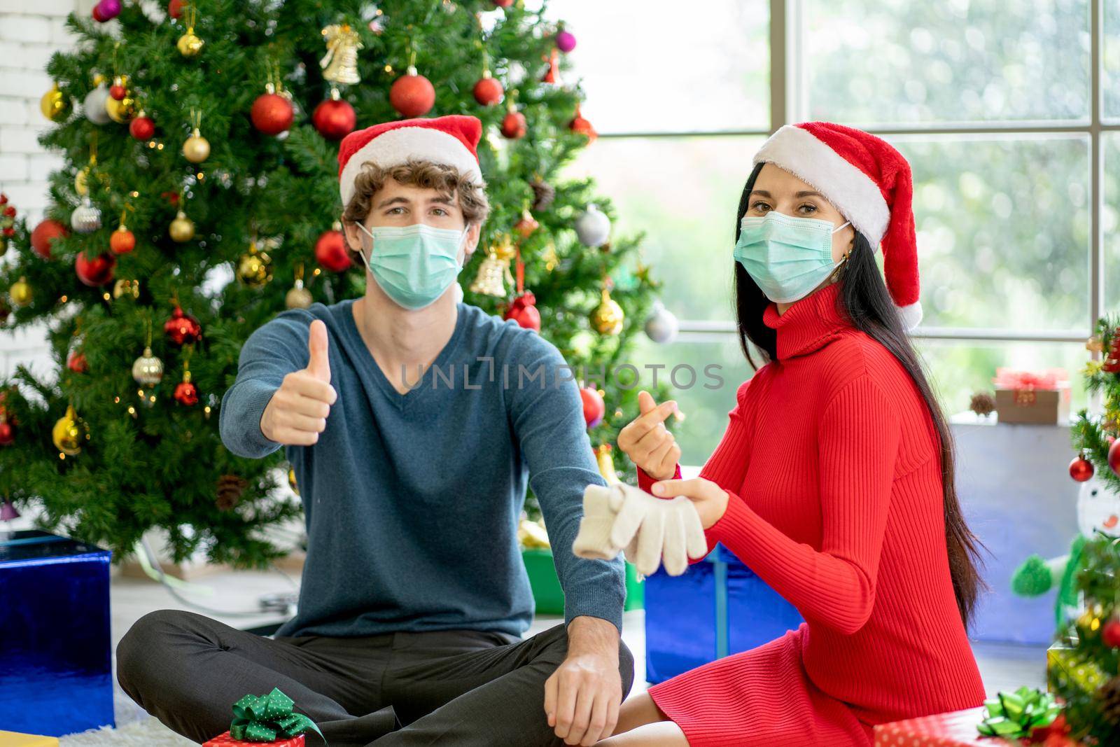Couple man and woman with hygiene mask show thumbs up and mini heart to camera with celebration of Christmas during pandemic of Covid-19 and stay at home with new normal lifestyle.
