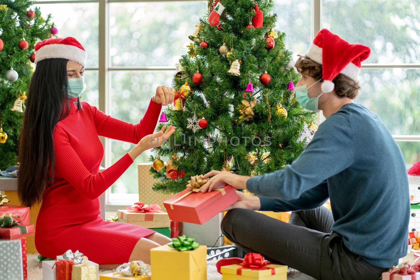 Couple man and woman with hygiene mask discuss and help to decorate Christmas tree together in room of their house. by nrradmin