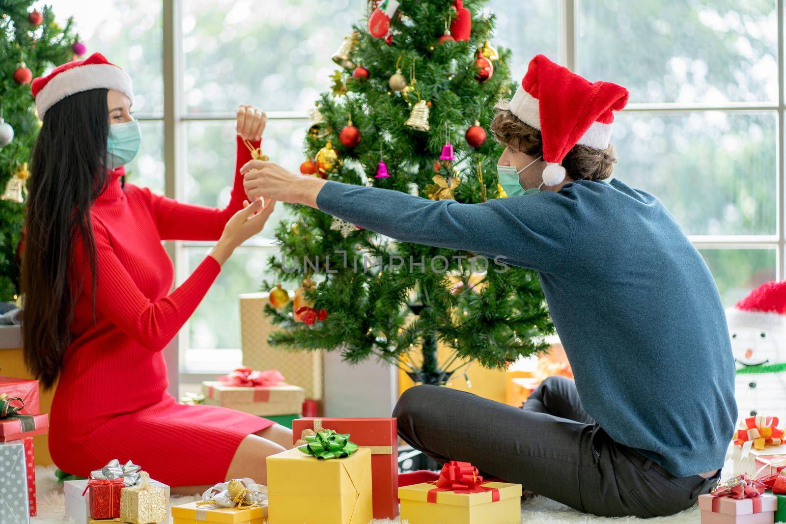 Couple man and woman with hygiene mask discuss and help to decorate Christmas tree together in room of their house. by nrradmin