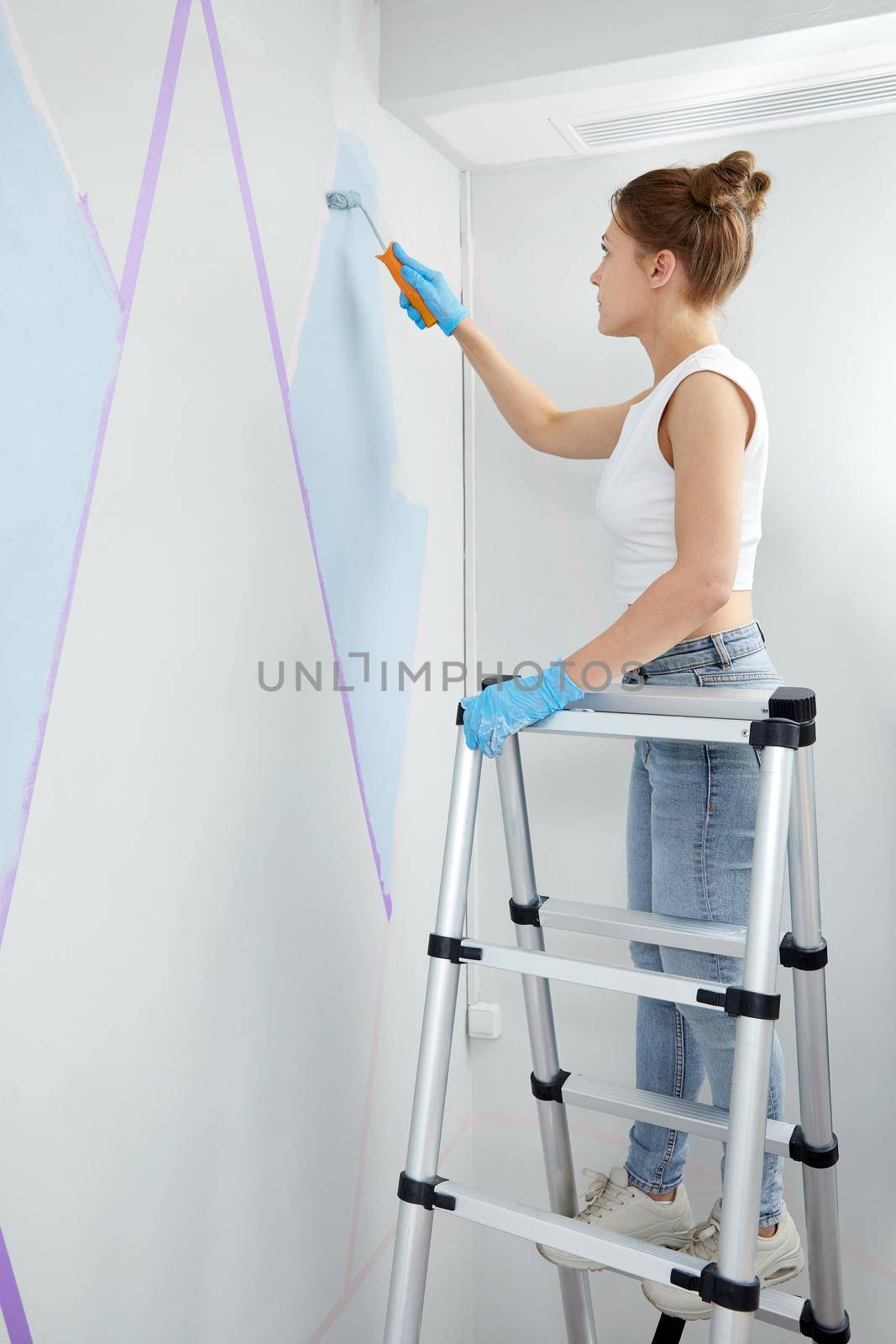 Young woman painting wall with paint roller and using masking tape while standing on ladder by Mariakray