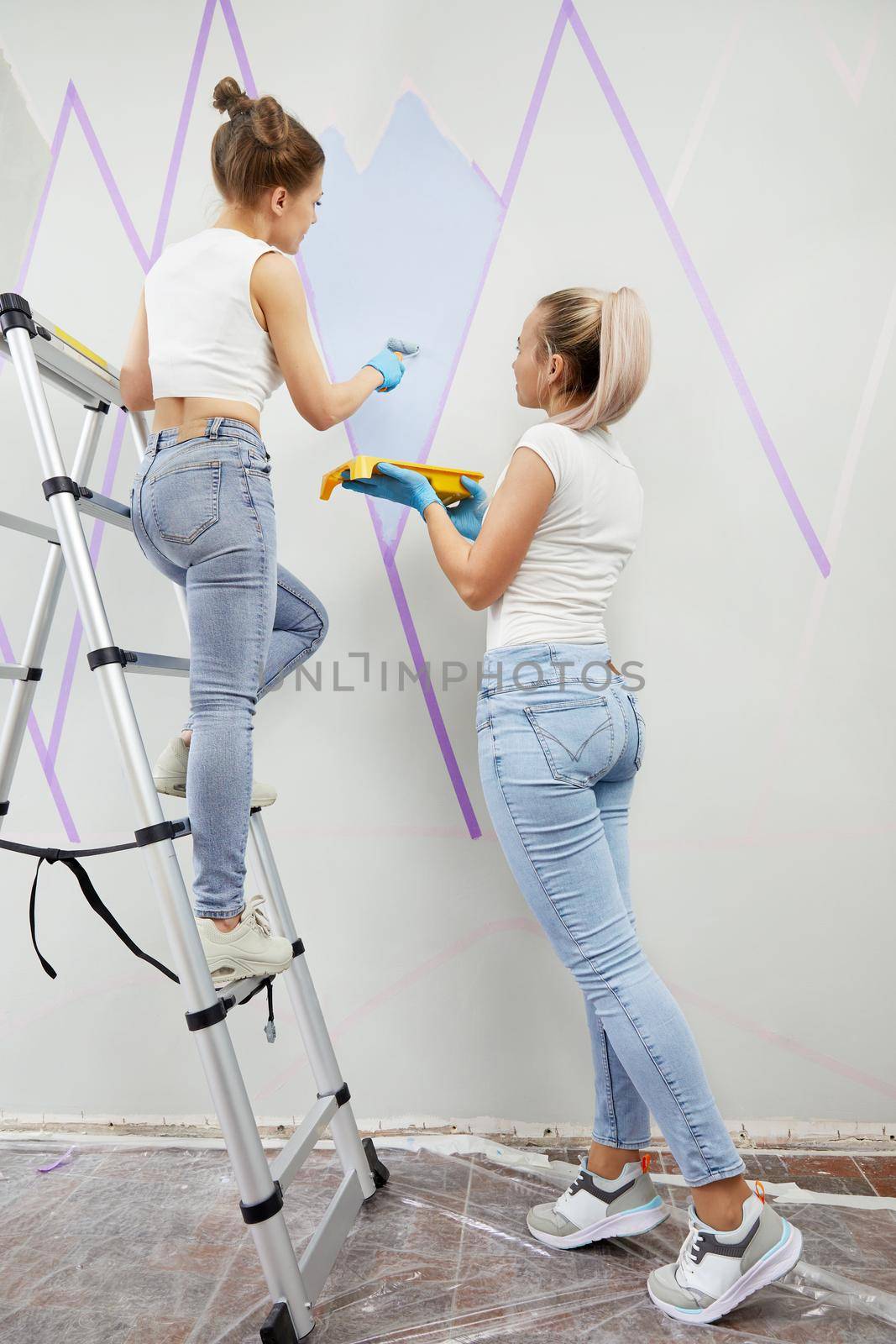 Two young women painting wall with paint roller and using masking tape while standing on a ladder by Mariakray