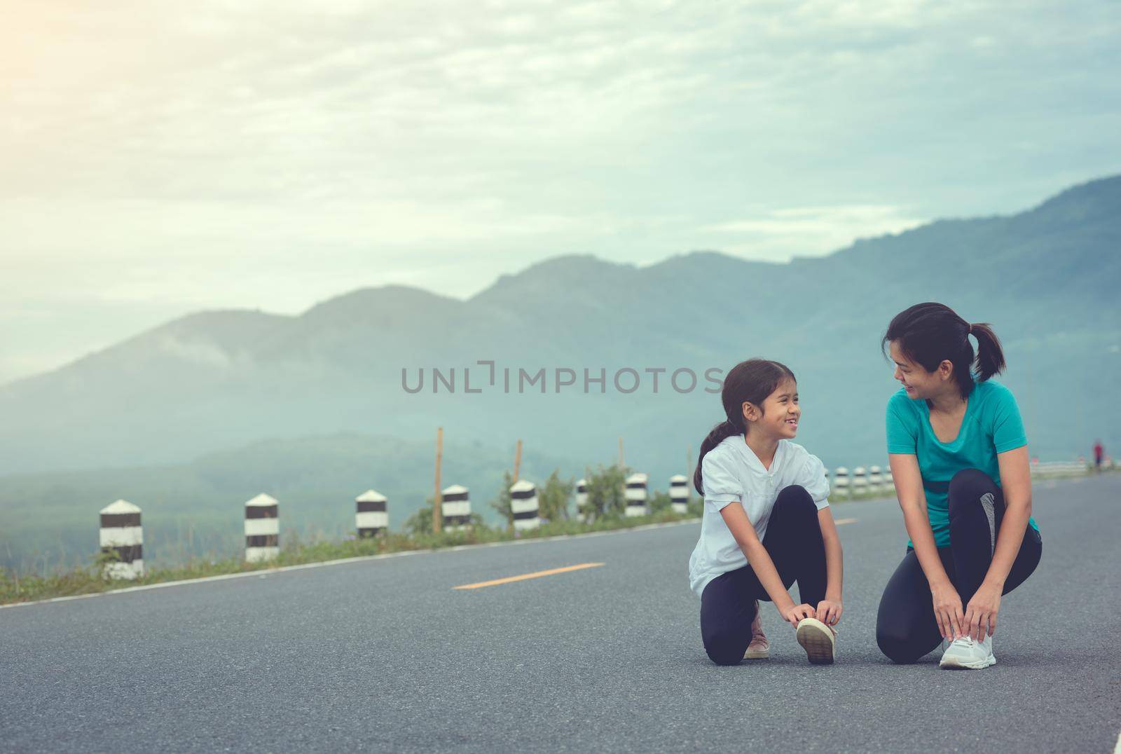 Mom and child are kneeling and tying shoelace. We getting ready for jogging outdoors the time during sunrise on dam road exercise. by thanumporn
