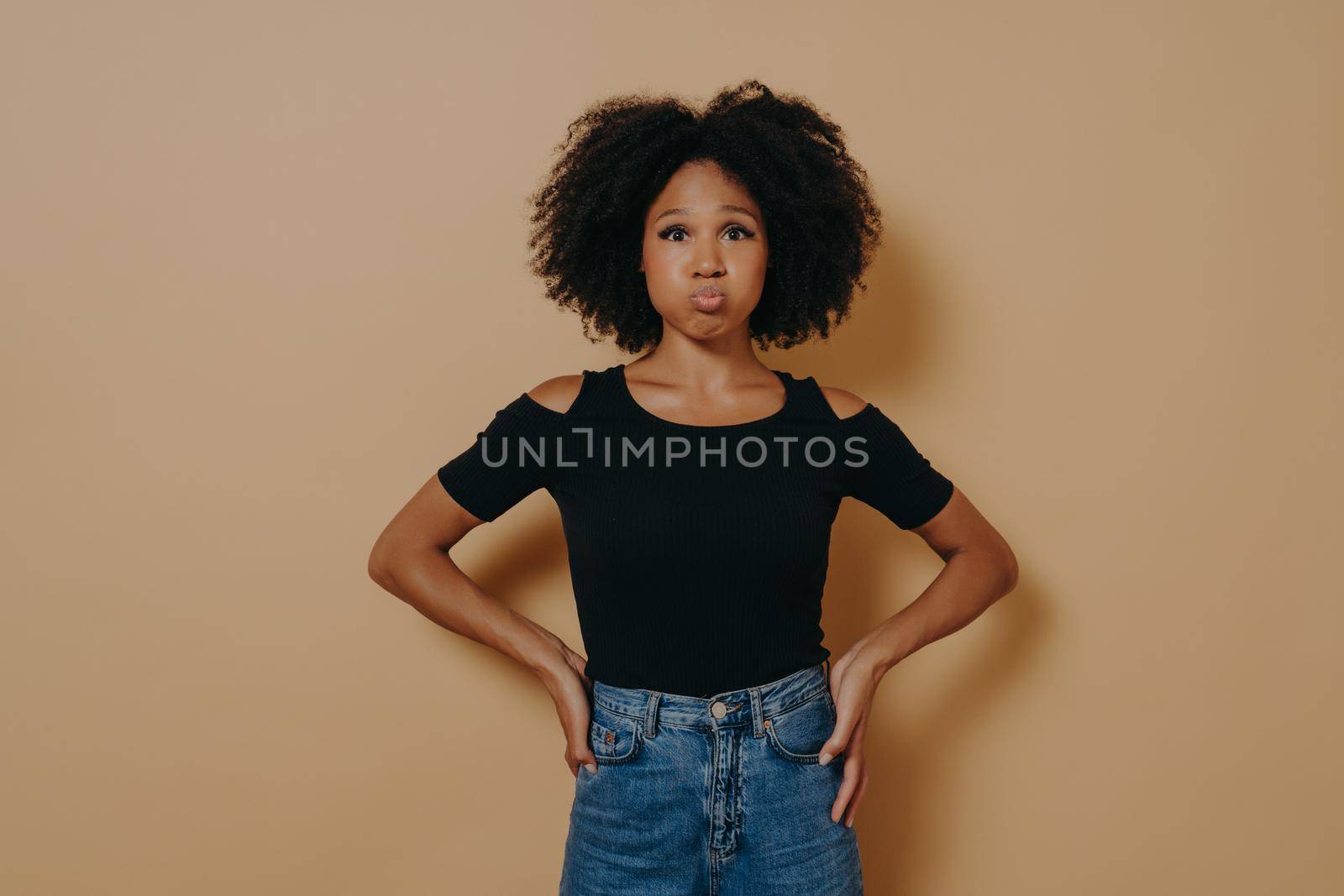 Funny carefree young dark skinned female in casual outfit blowing puffing out her cheeks and having fun indoors against nude studio wall background. People and fun concept, face expressions
