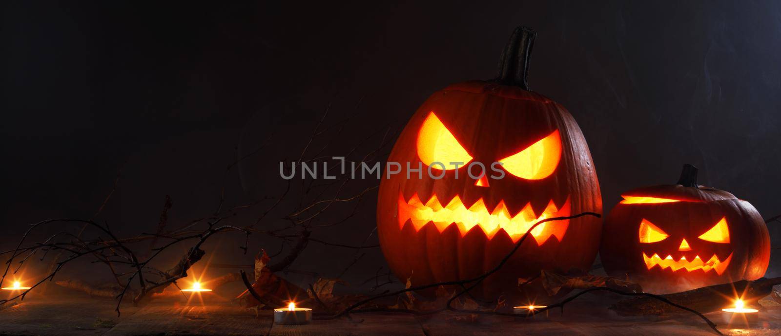 Halloween two luminous evil scary pumpkins, jack-lantern, with candles, leaves and eerie branches on wooden dark background