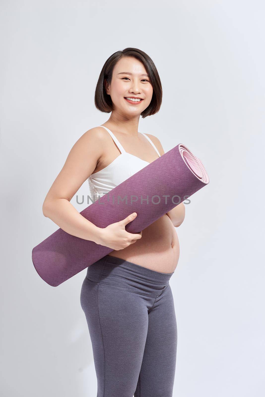 Joyful pregnant woman holding yoga mat smiling to camera posing over white background. Pregnant lifestyle and fitness. by makidotvn