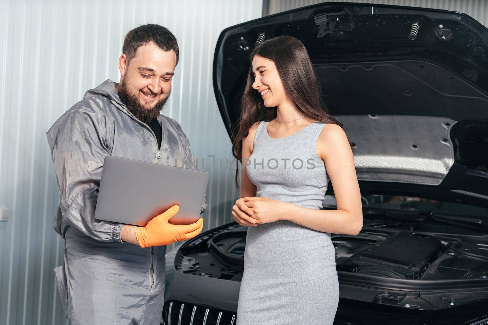 Car mechanic communicating with a customer while using laptop and examining vehicle breakdown at auto repair shop