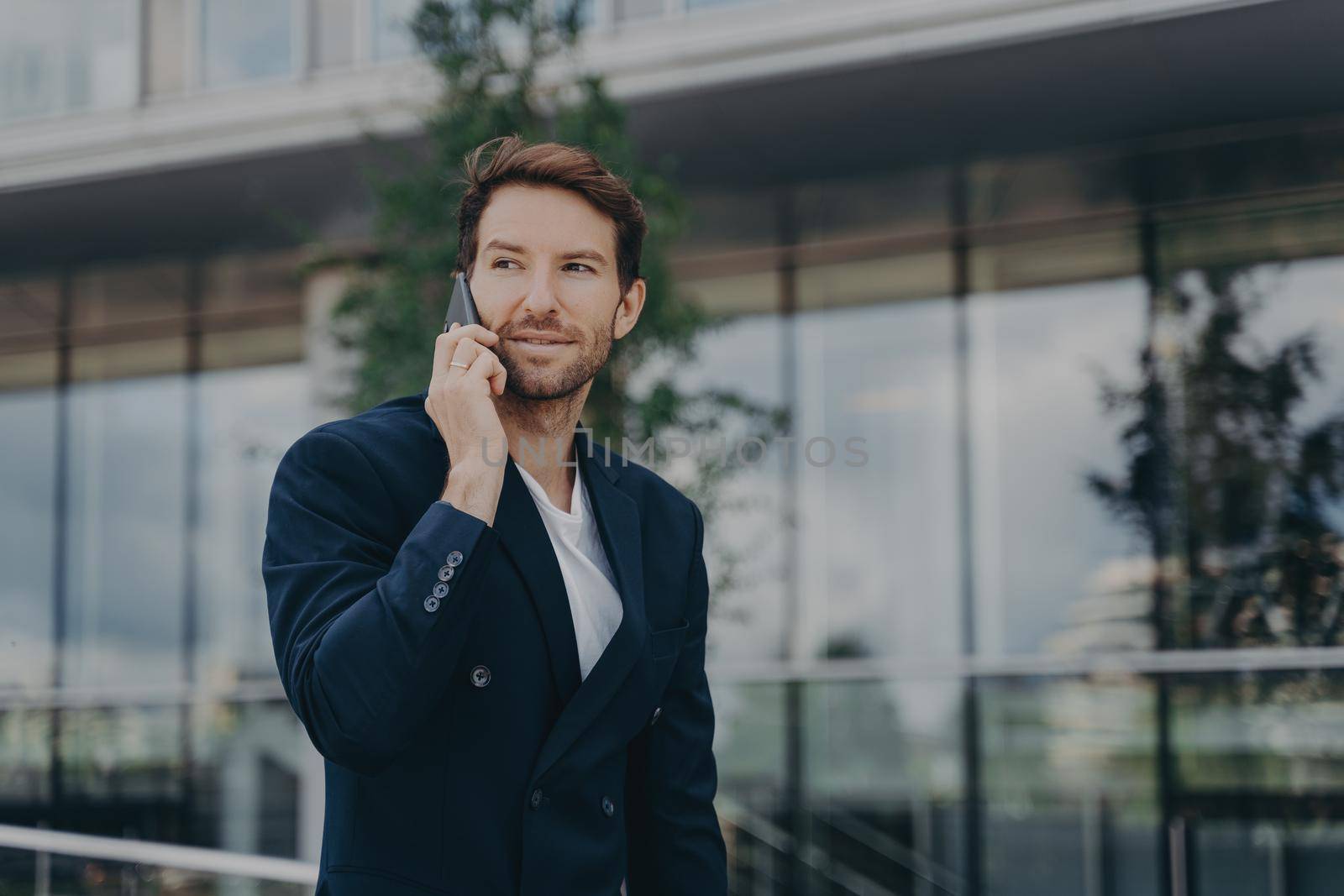 Male entrepreneur talks via phone solves urgent problems poses near business center dressed in black suit uses modern technologies for communication looks away thoughtfully. Technology work lifestyle