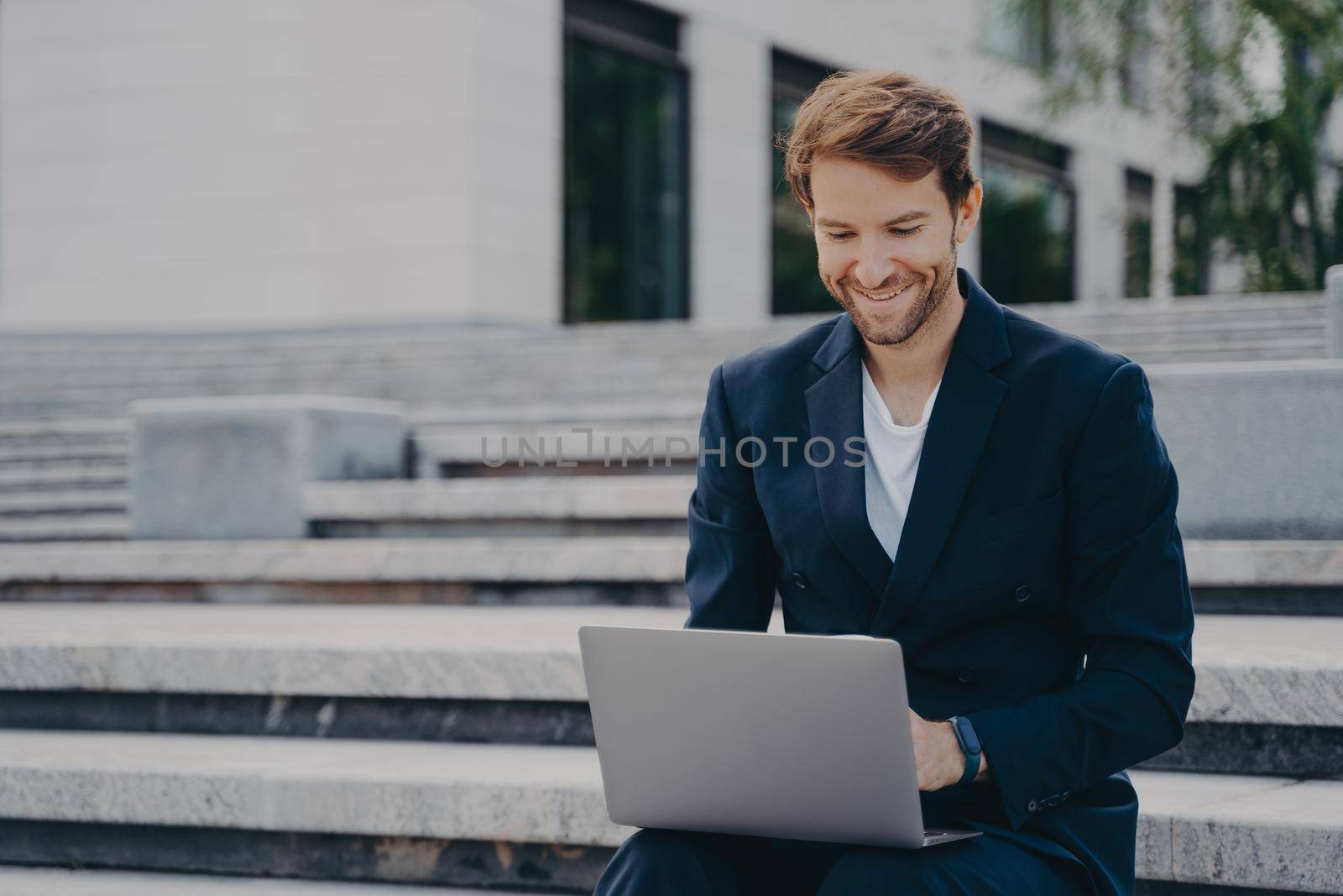 Young businessman sits on steps uses laptop organizes meeting online with investors has happy expression uses free internet poses outdoor dressed formally. Corporate director analyzes web information