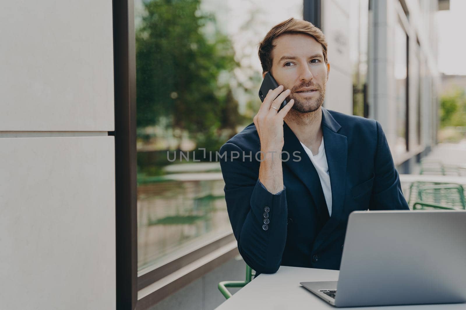 Outdoor shot of successful man executive worker calls someone via smartphone consults customer by phone sits at table in cafe waits for order uses laptop for work online. Manager holds interview