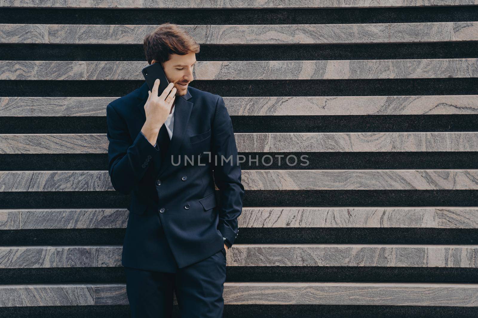 Businessman in elegant black clothes phones to partner focused down keeps smartphone near ear poses near marble wall copy space for your advertisement. People technology and bussines concept