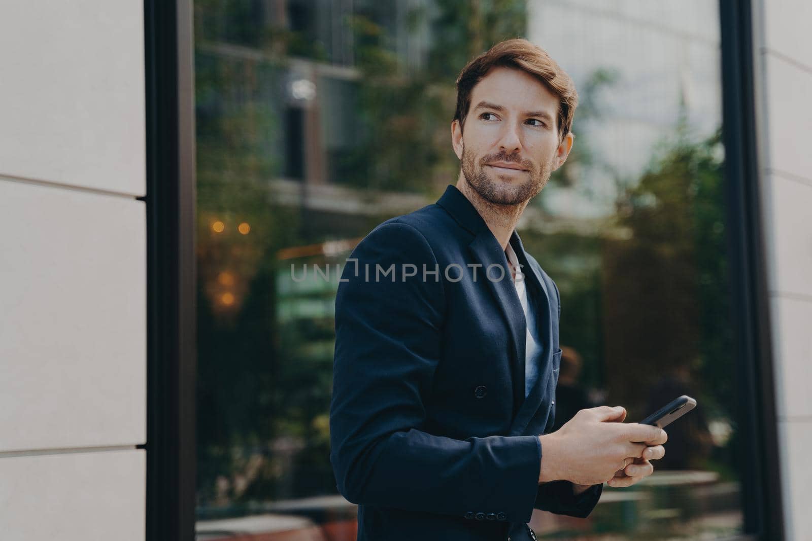 Successful handsome businessman with stubble messaging his employer about project details, holding smartphone in his hands while standing next to building with big window reflecting urban landscape