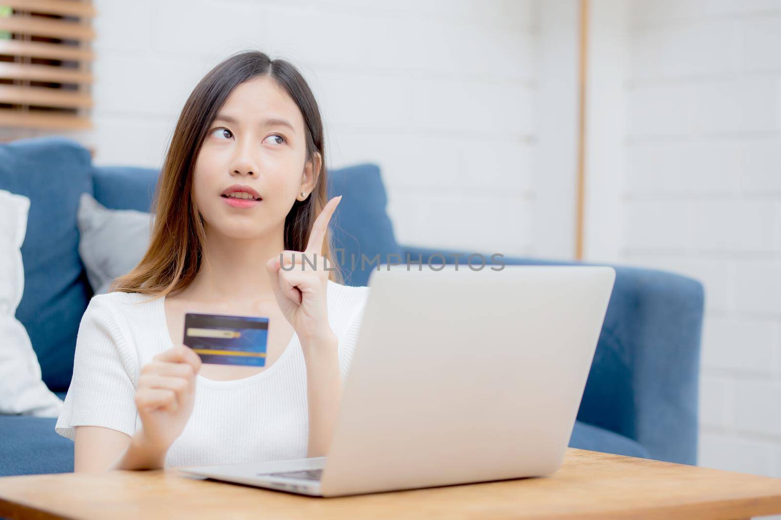 Young asian woman sitting thinking idea using credit card with laptop computer on couch, girl shopping online for buy and payment with notebook on sofa, finance and debit, lifestyle concept.