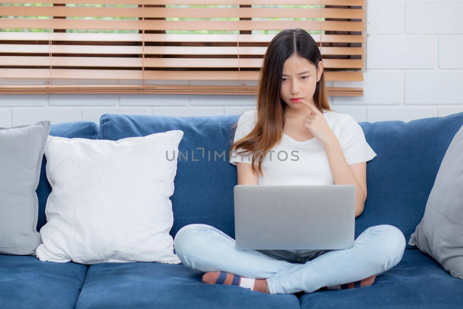 Beautiful young asian woman working on laptop computer and thinking idea on sofa at home, freelance girl sitting on couch using notebook to internet at living room, one person, lifestyle concept.