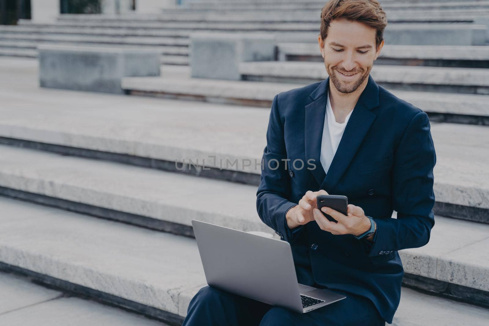 Young happy male entrepreneur in stylish suit using smartphone, reading messages from clients and smiling, using laptop while working online remotely, sitting on stairs outside