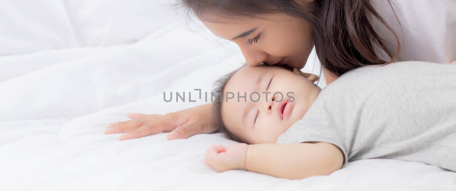 Young asian mother kiss cheek of little baby girl with tender on bed in the bedroom, mom love newborn and care, mother with expression with child together, parent and daughter, family concept.