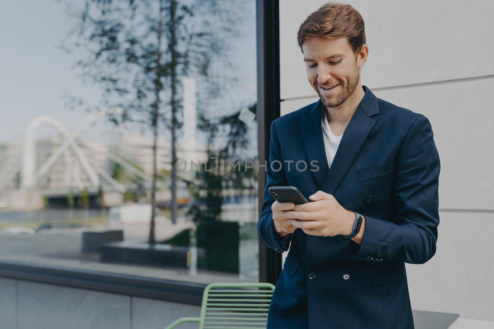 Pleased handsome male CEO or director holds mobile phone waits for call scrolls social networks while waiting for partner near outdoor cafe wastes time online dressed in elegant formal suit.