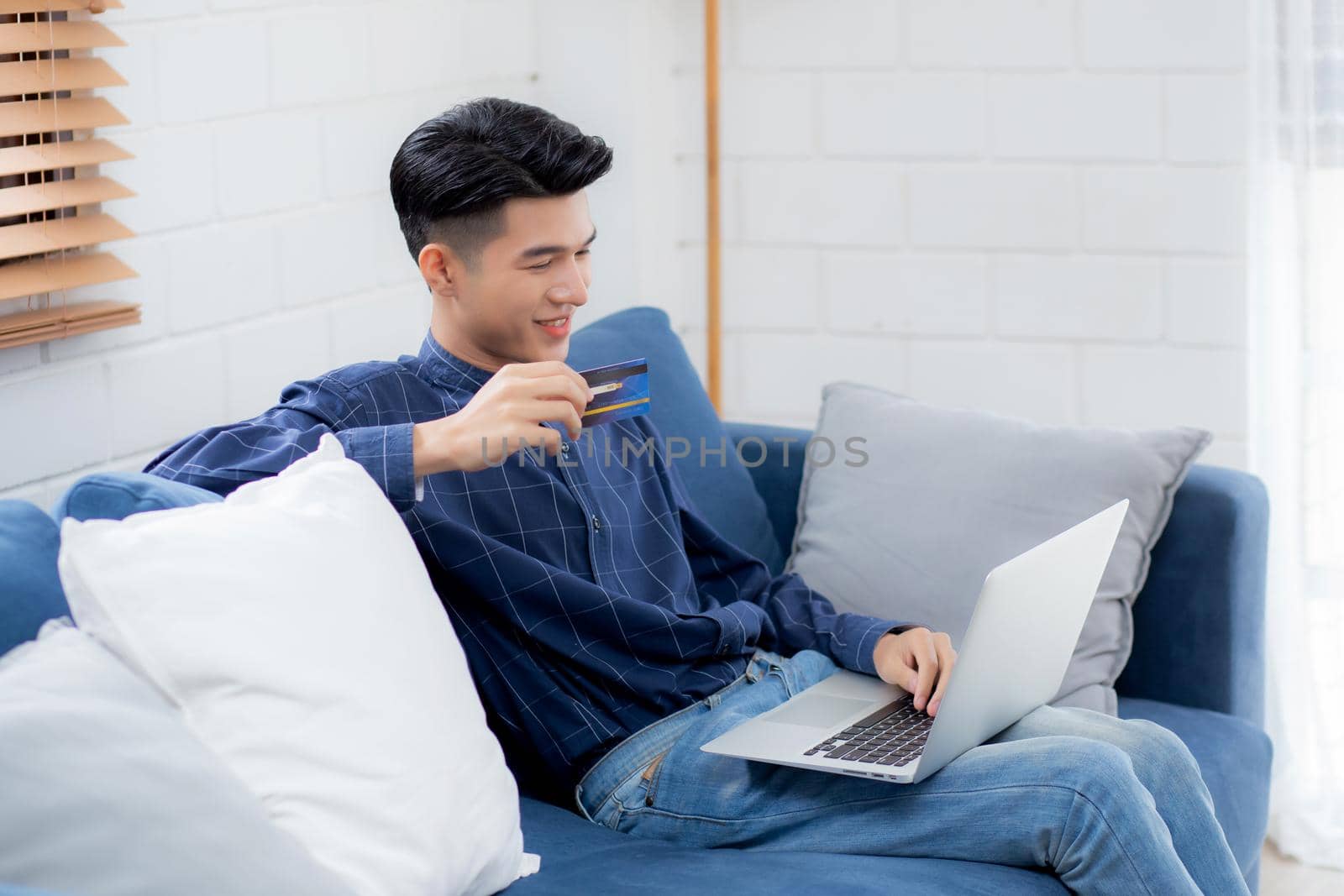 Young asian man sit on sofa using laptop computer shopping online with credit card buying to internet, happy male payment with business on couch, purchase and payment, business concept.