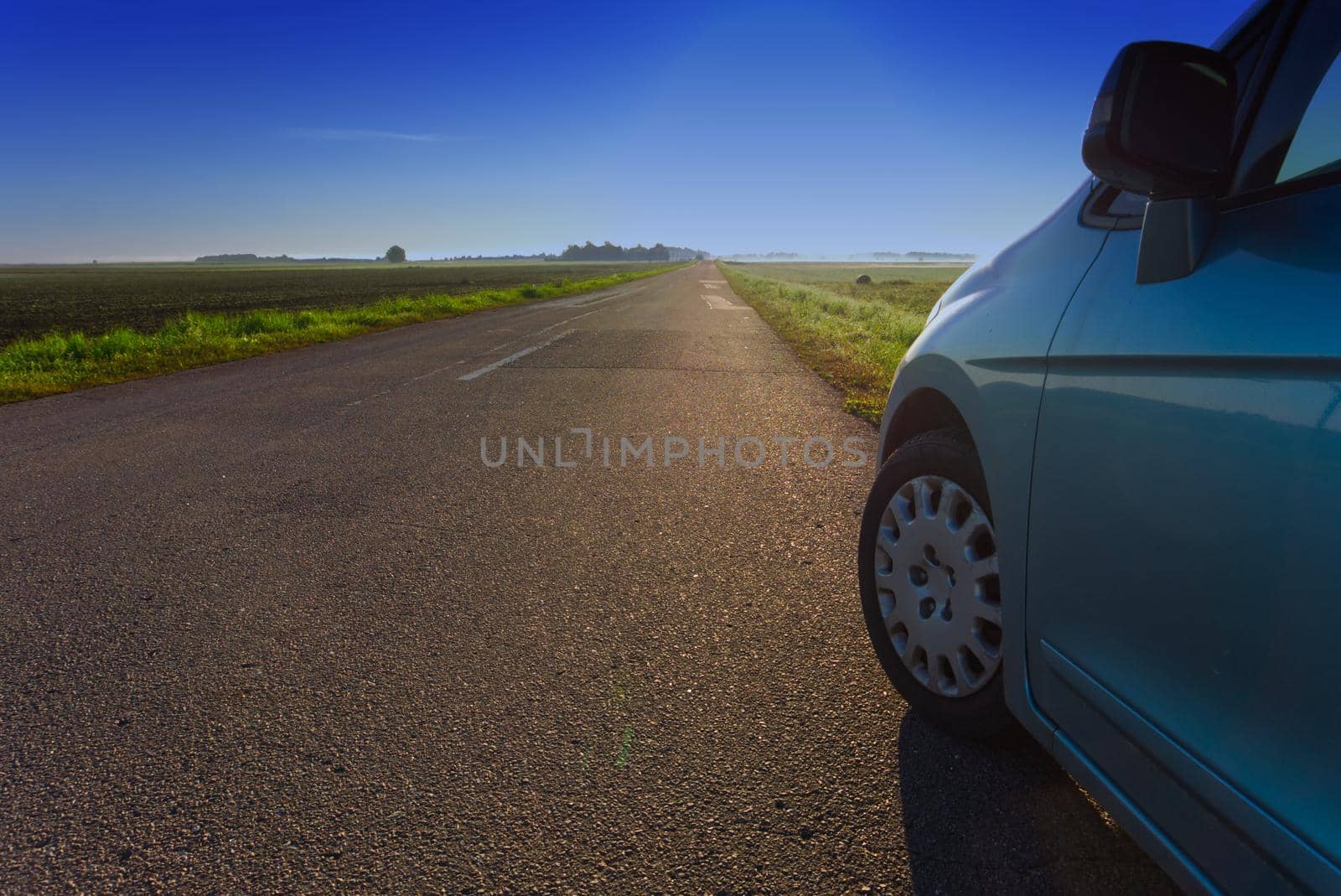 Close-up view of the side of a blue car parked at the edge of an empty country road through a green field towards a clear blue sky in Spring or Summer