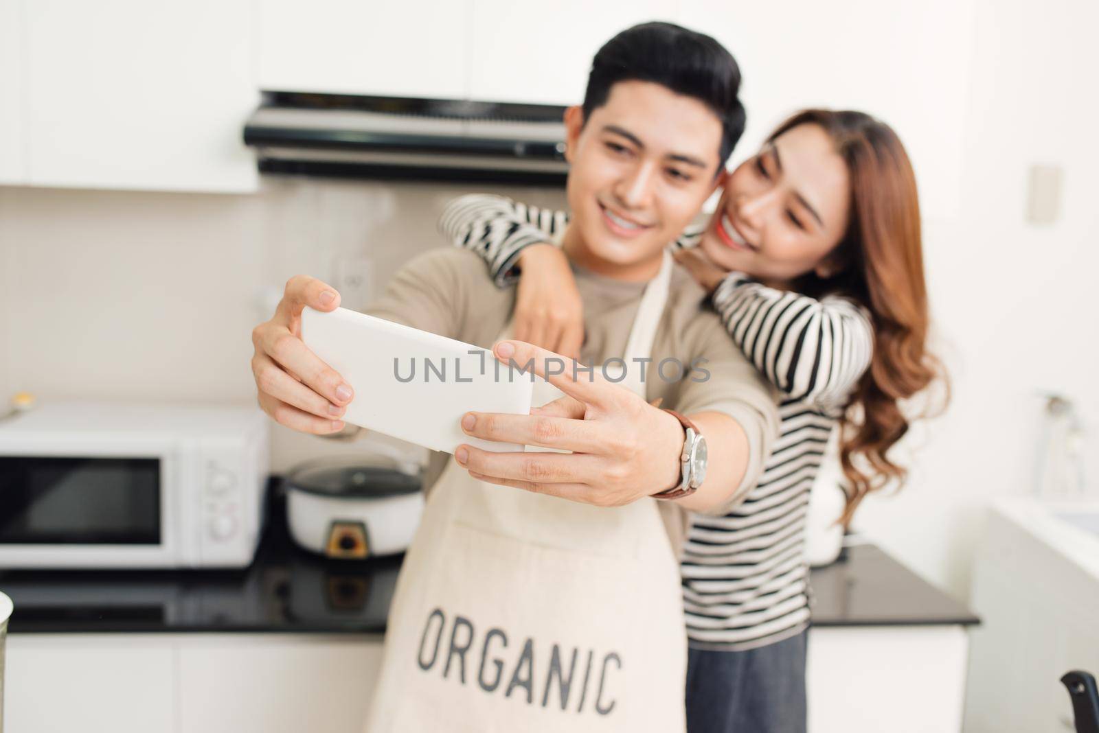 Happy asian couple smiling and cooking in the kitchen doing selfie using a smart phone by makidotvn