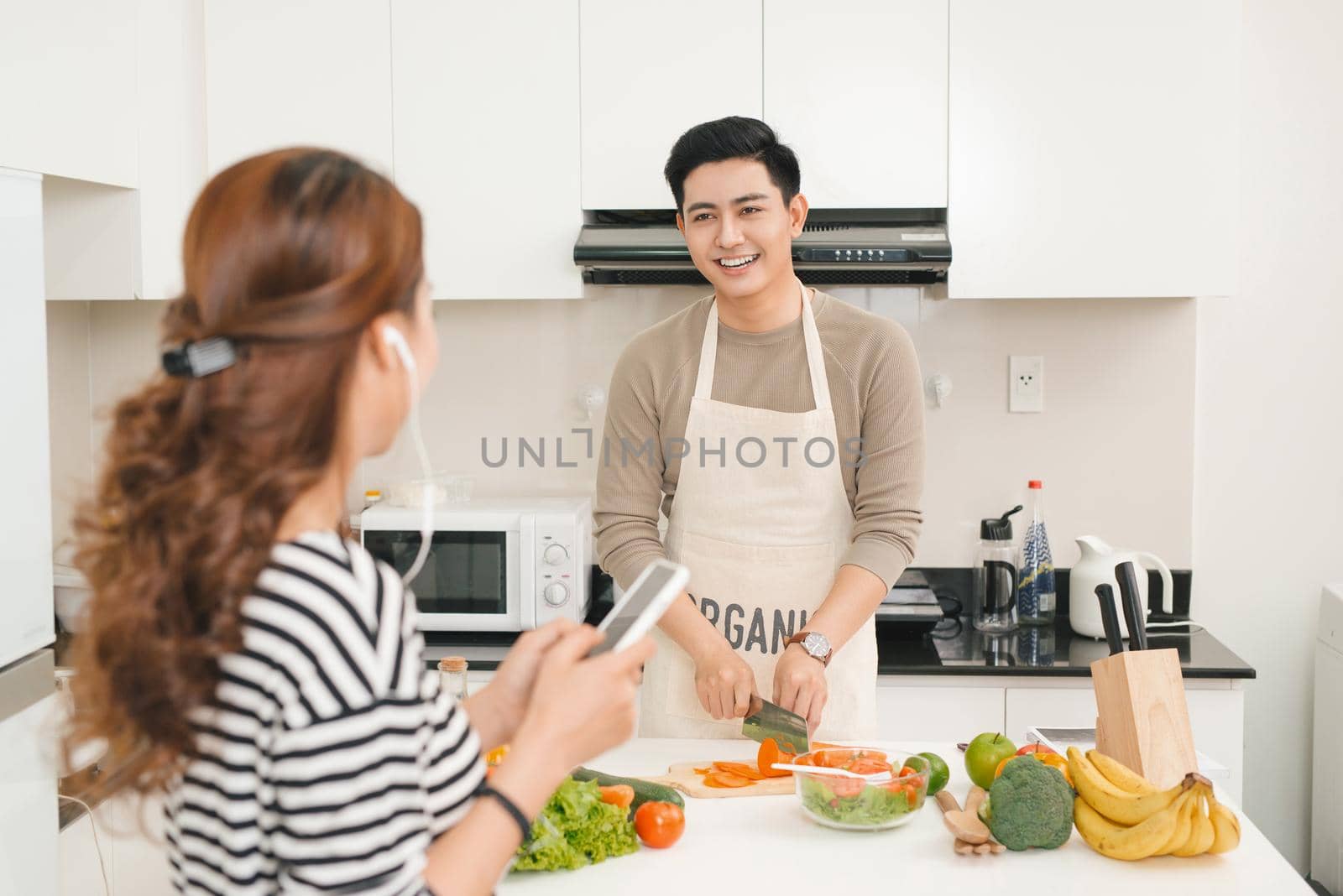 Woman using mobile phone in kitchen as boyfriend prepares meal by makidotvn
