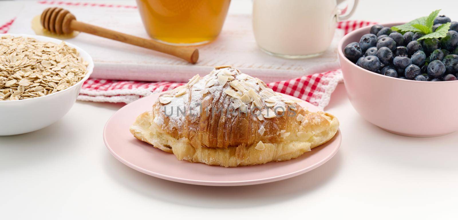 baked croissant sprinkled with powdered sugar, blueberries and oatmeal in a ceramic plate on a white table, breakfast