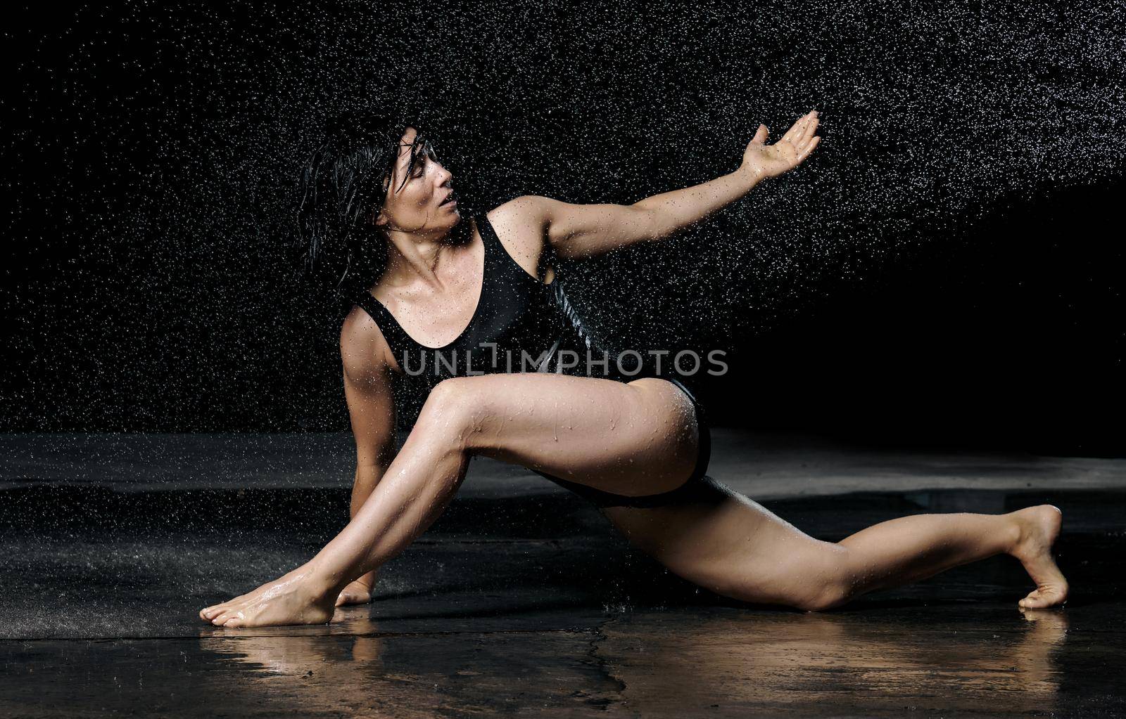 woman with black hair lies on the ground under raindrops on a black background. Woman dressed in black bodysuit by ndanko