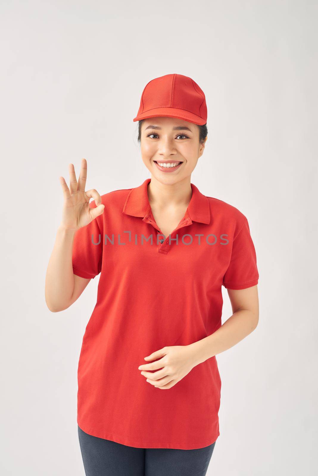 Happy young girl courier, wearing cap uniform looking at camera showing ok sign recommend fast delivery service on white background. by makidotvn