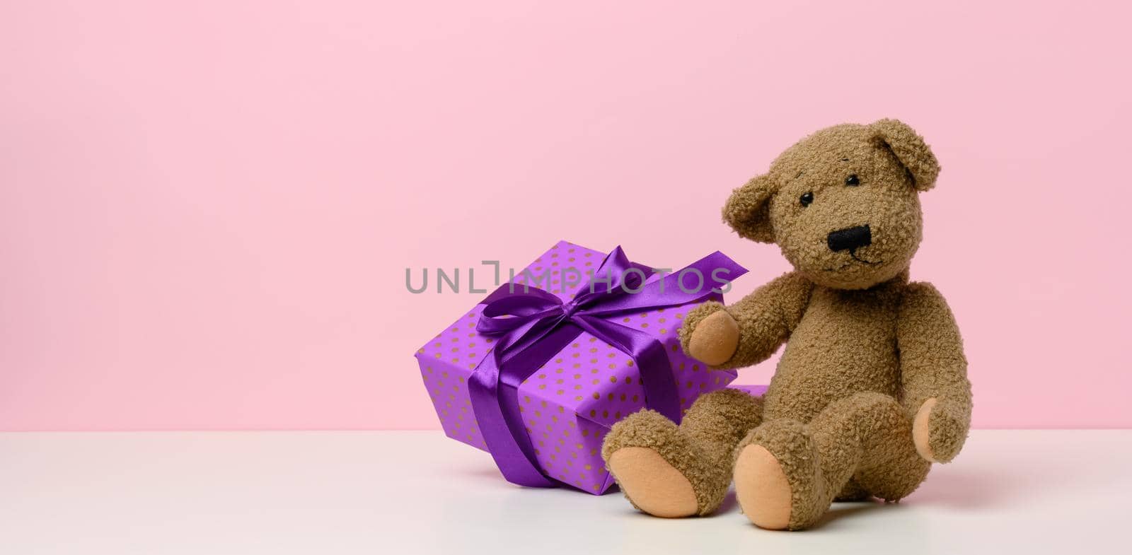 cute brown teddy bear holding a box wrapped in paper and red silk ribbon on white table. Prize and congratulations, by ndanko