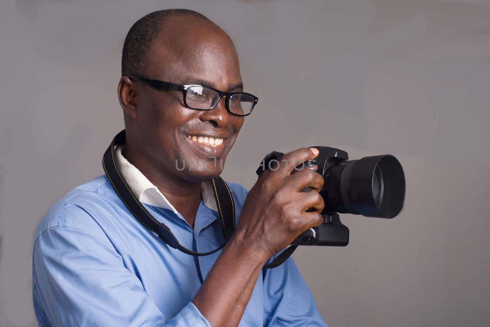 African man standing in blue shirt on gray background looking ahead with camera in hand.