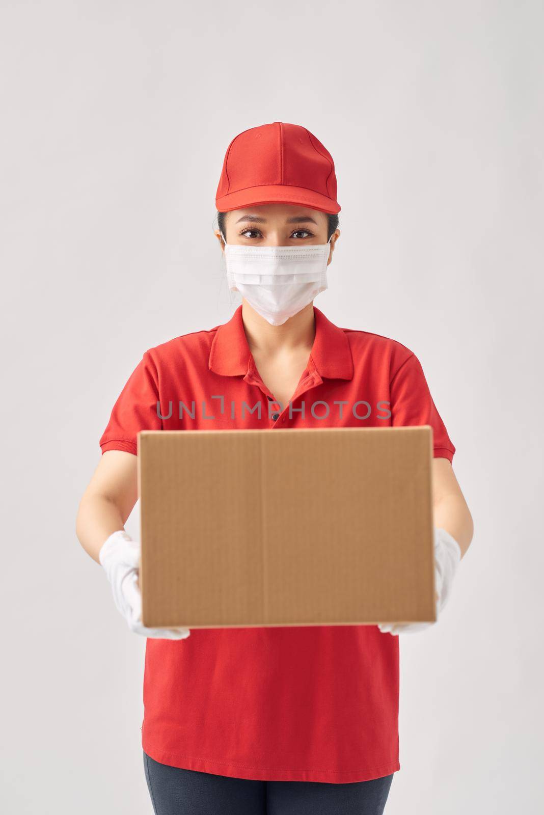 A uniformed courier wearing medical rubber gloves and a medical mask holds a paper box of food on a white background. 