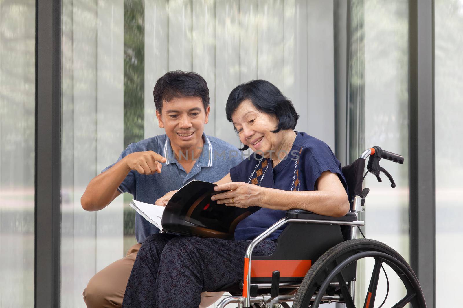 Elderly woman reading a book on wheelchair with her son take care.