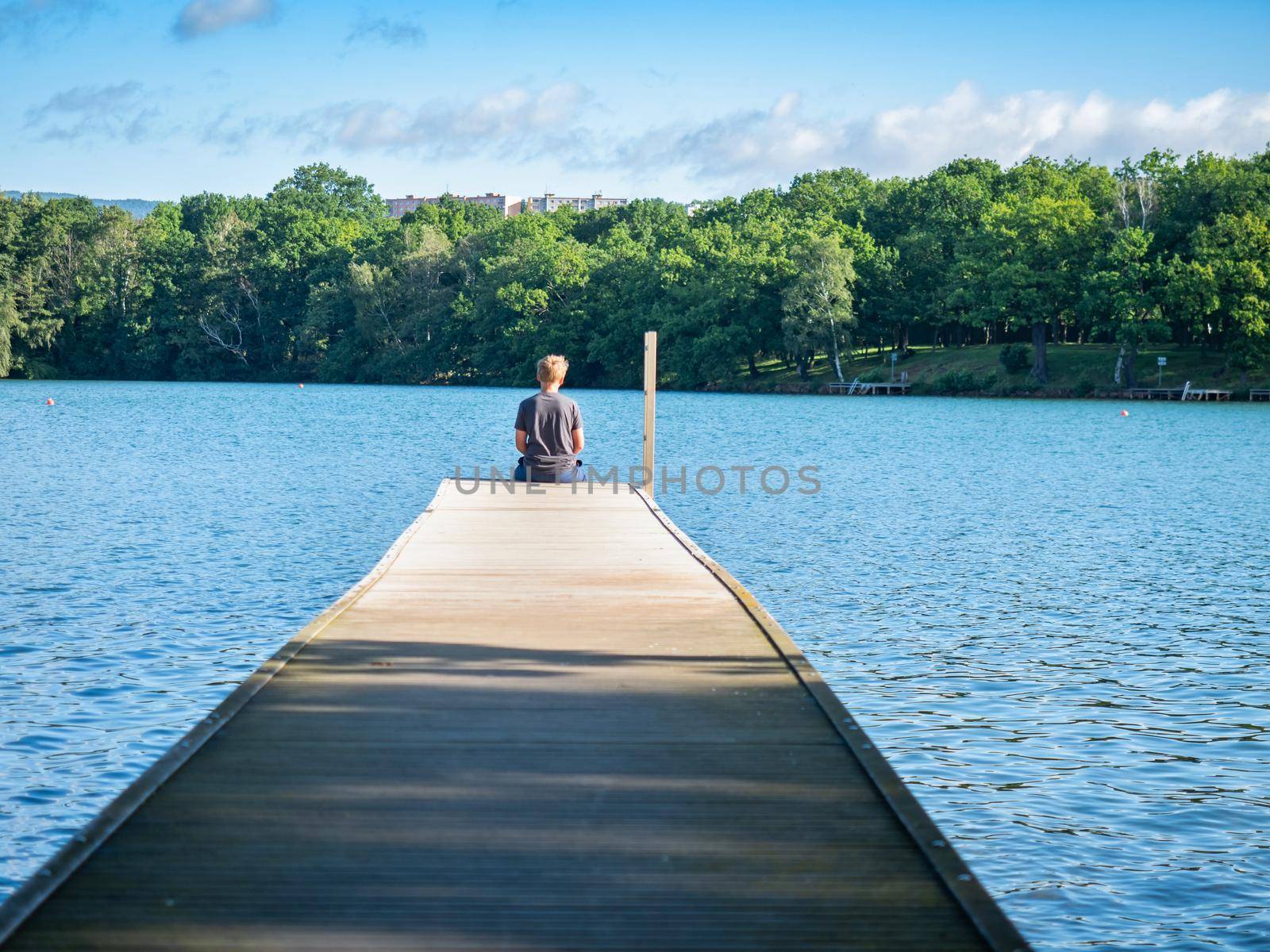Blond hair boy sits on the lake mole and looks at the water by rdonar2