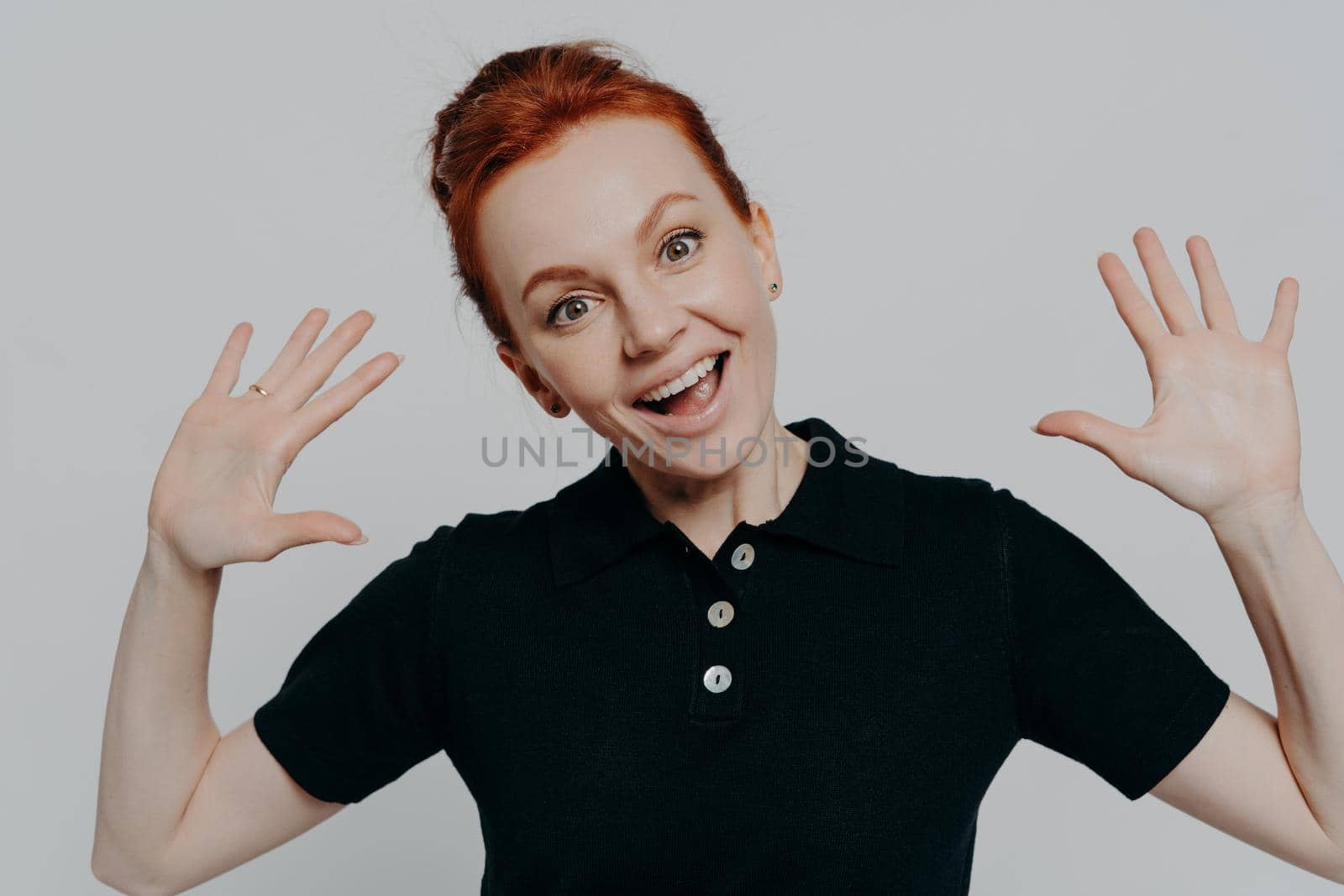 Portrait of young amused redhead woman raising palms and looking at camera with cheerful expression, excited ginger female seeing something funny, wearing black t-shirt, isolated on grey background