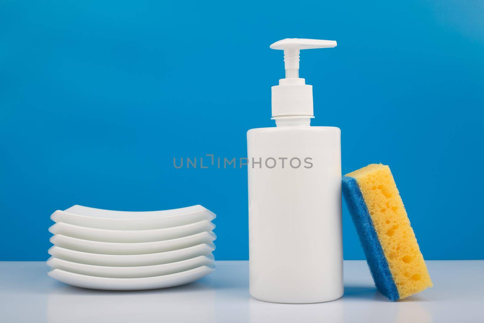 Dishwashing detergent in white bottle, pile of saucers and cleaning sponge against blue background by Senorina_Irina