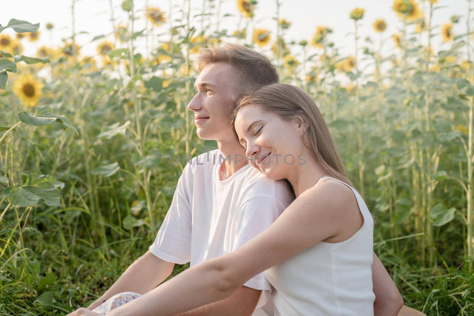 Young couple having picnic on sunflower field at sunset, woman lying on boyfriend shoulder by Desperada