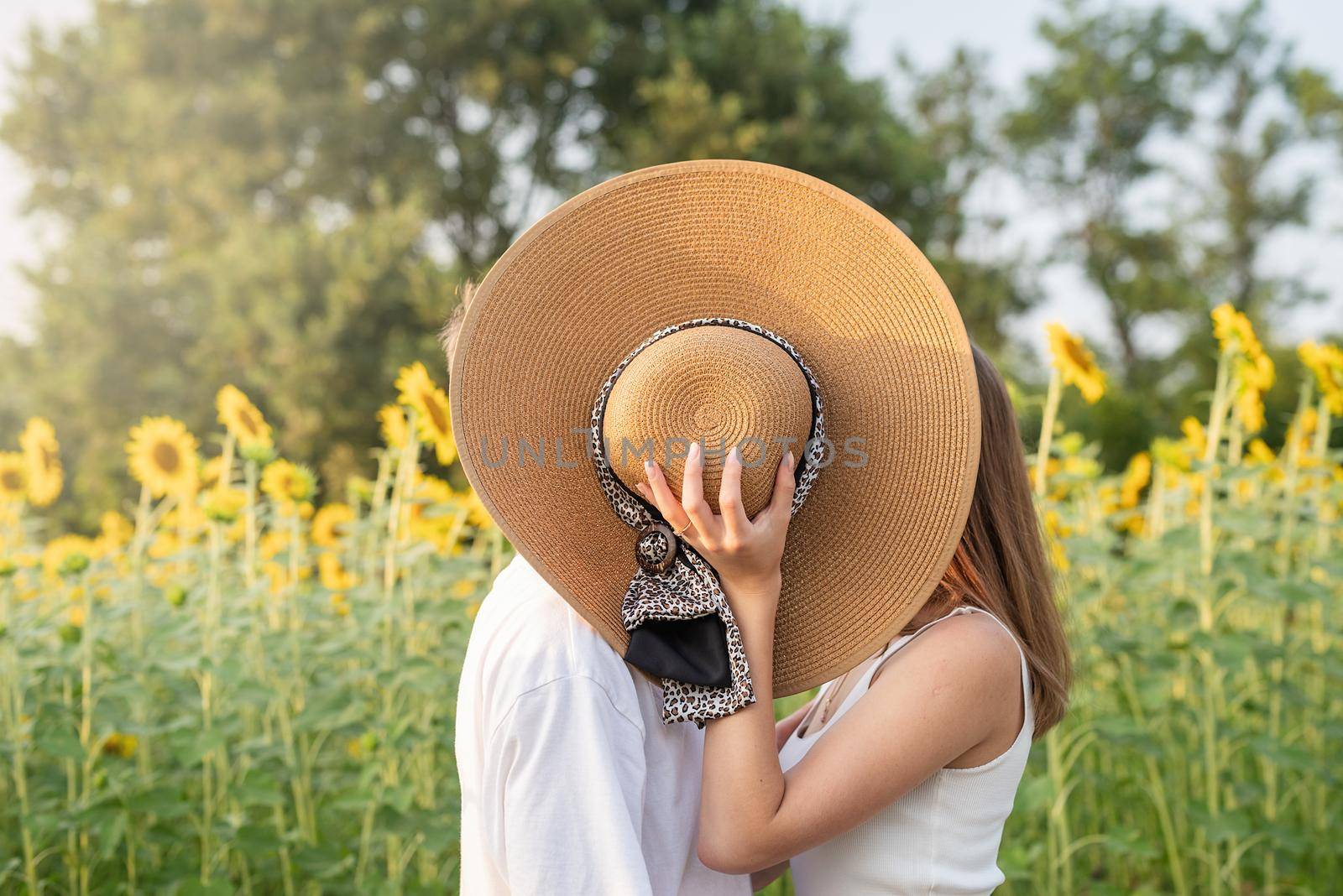 Young happy couple kissing on a picnic blanket, covering their faces with a summer hat by Desperada