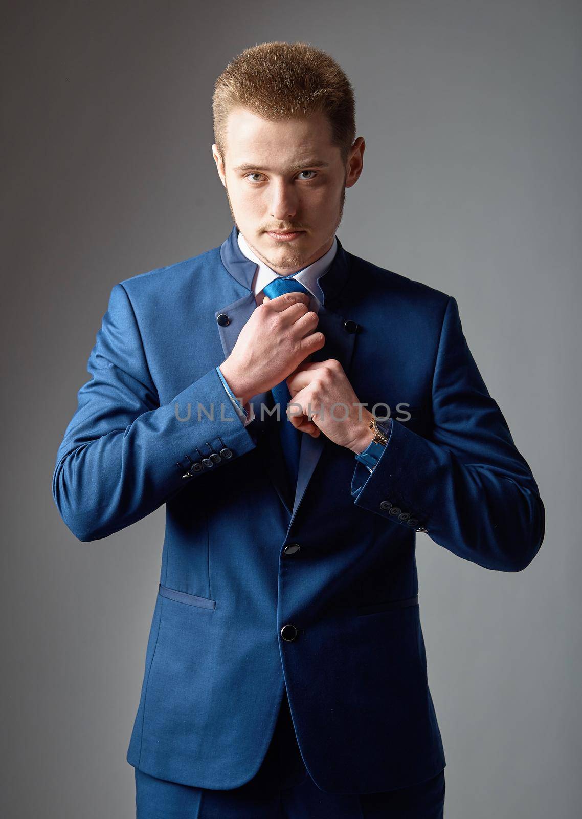 Handsome and self-confident. Handsome young man in formalwear adjusting his necktie and looking at camera while standing against grey background