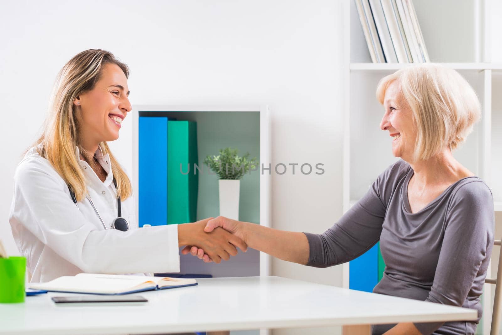 Female doctor and happy senior woman patient shaking hands in doctor's office.