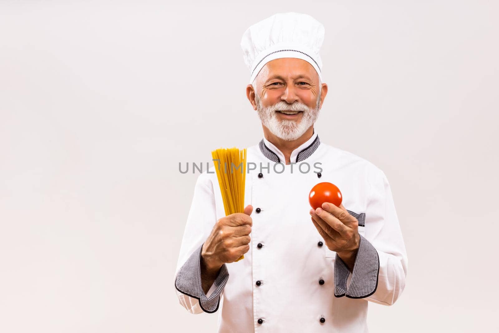 Portrait of senior chef holding tomato and spaghetti on gray background.