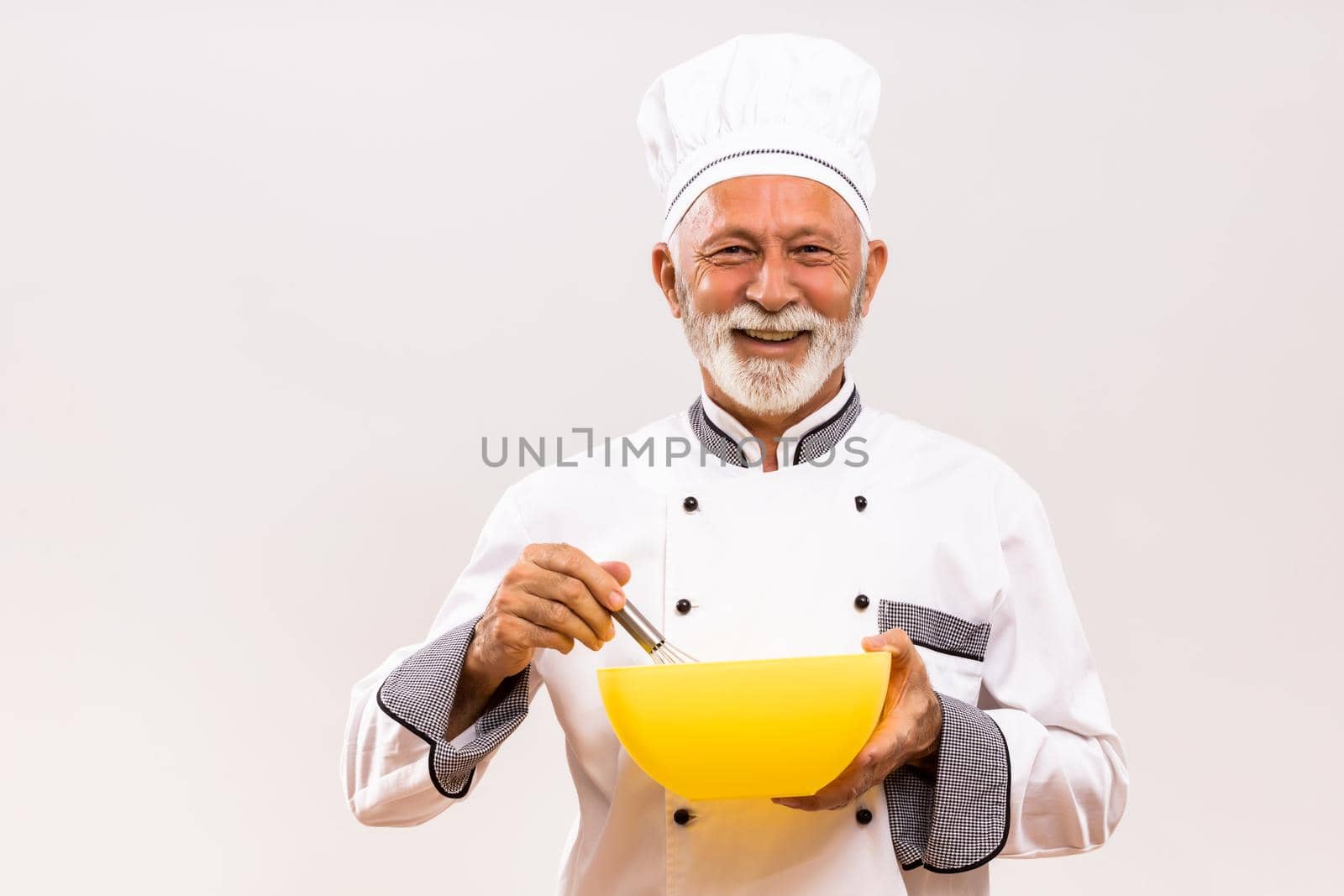 Senior chef holding bowl and wire whisk on gray background.