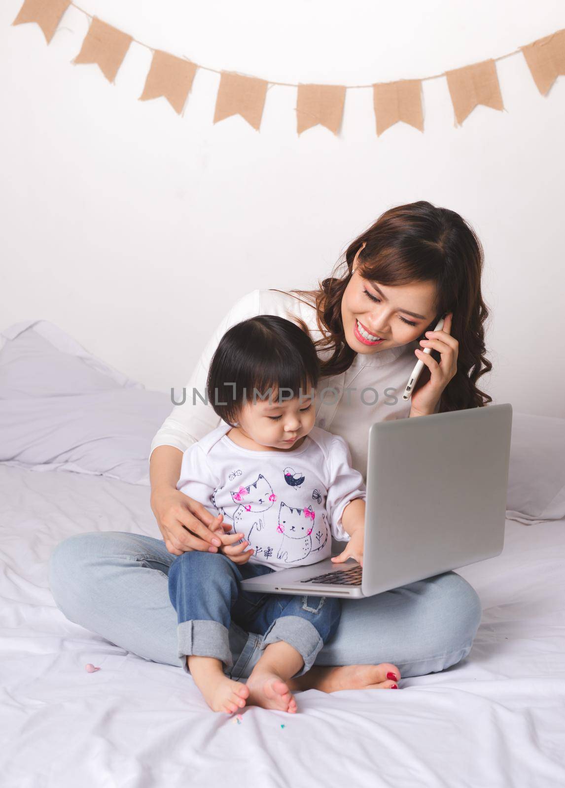 Asian lady in classic suit is talking on the mobile phone and working on laptop at home with her baby girl. by makidotvn