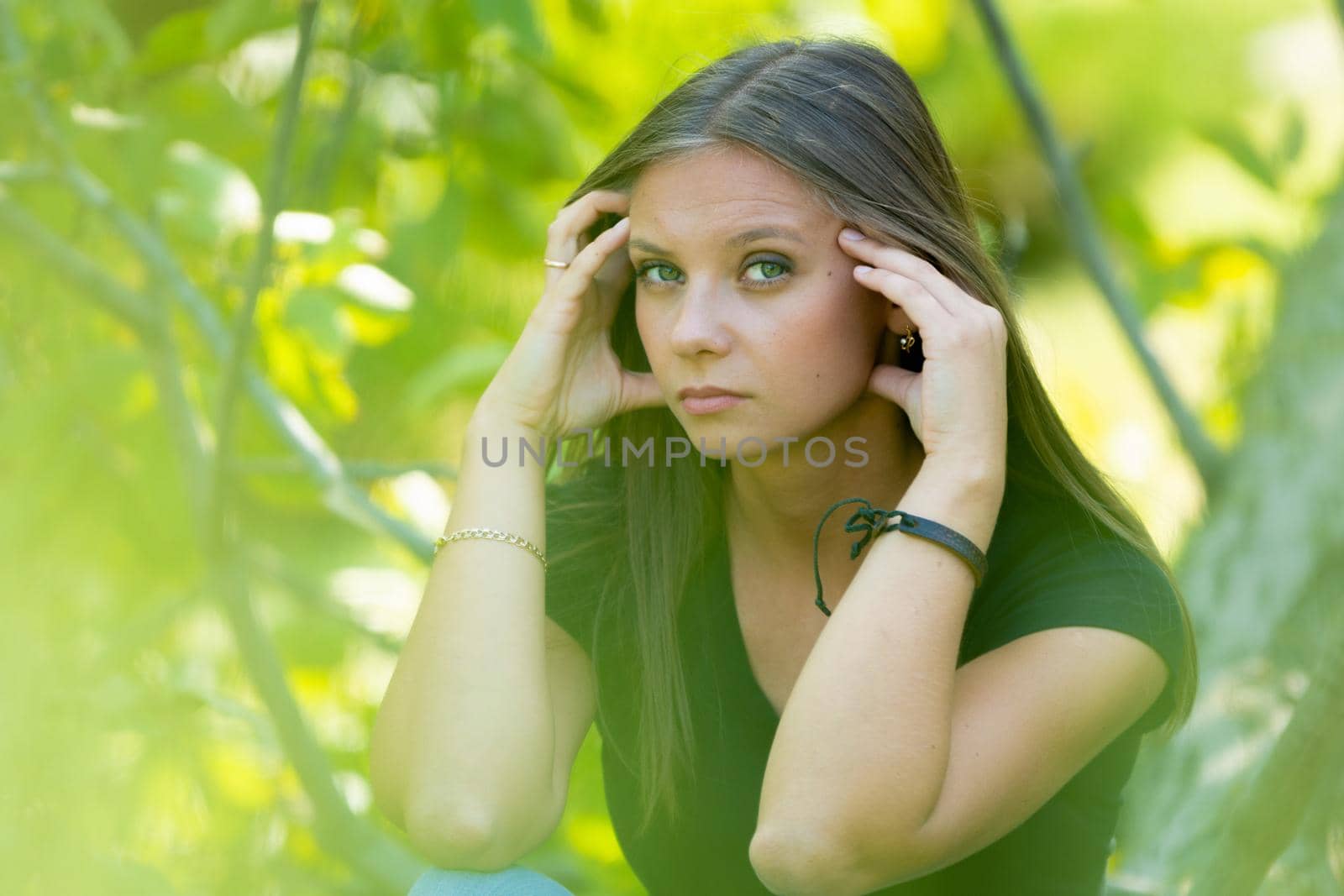 A sad girl sits on a background of foliage, holds her head with her hands and looks sadly into the frame