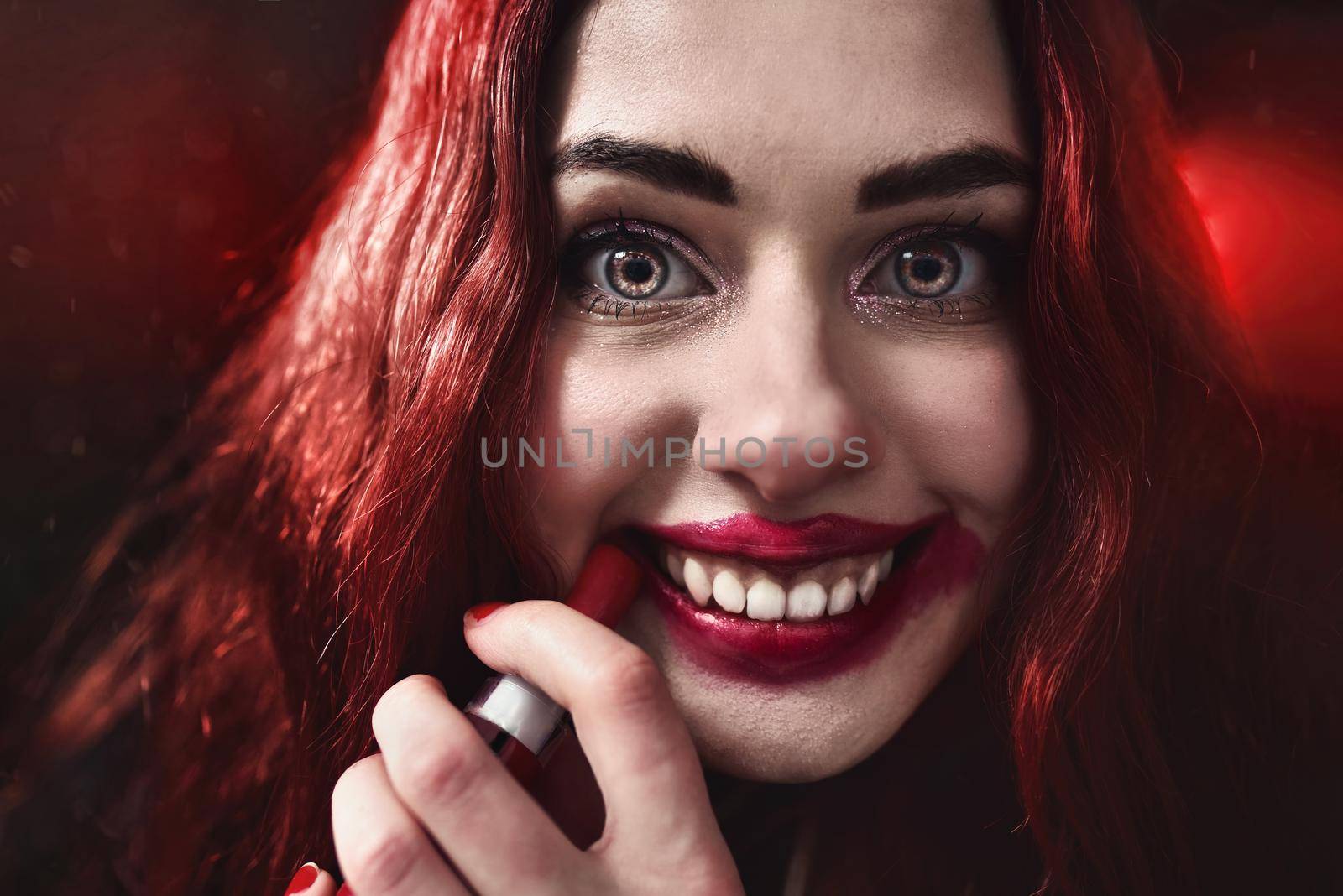 Halloween Time. Portrait of crazy-looking horror woman with red hair she is smearing red lipstick on her face, horror concept. Fear and nightmare