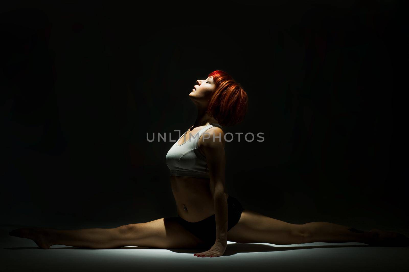 Fit red-haired woman wearing sports clothing doing the splits in the spotlight