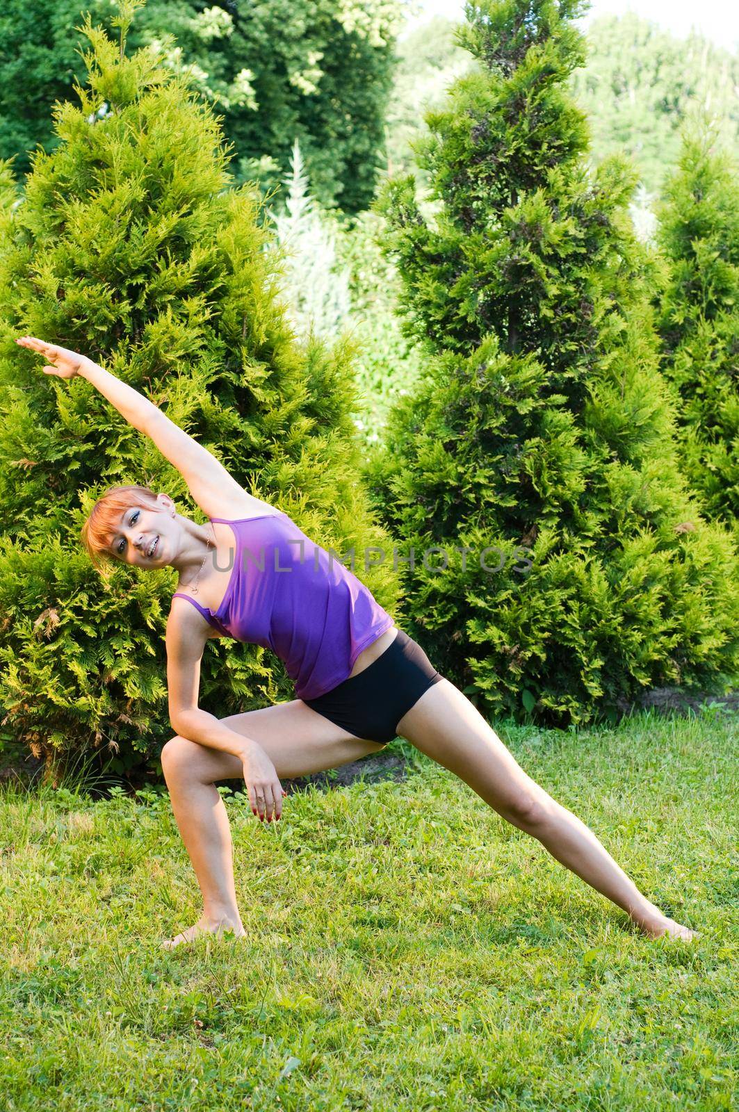 Beautiful red woman doing fitness or yoga exercises outdoors in a green park