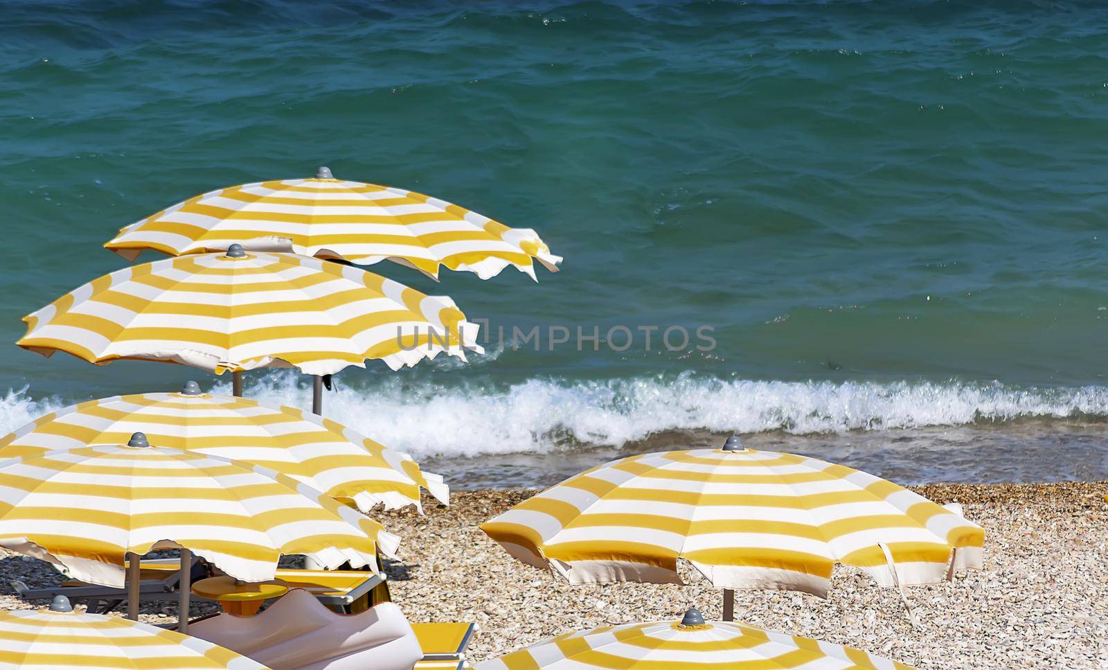 Yellow and white striped umbrellas open in a pebbly beach during a windy summer day. Sea waves on the background. Holidays and relaxation