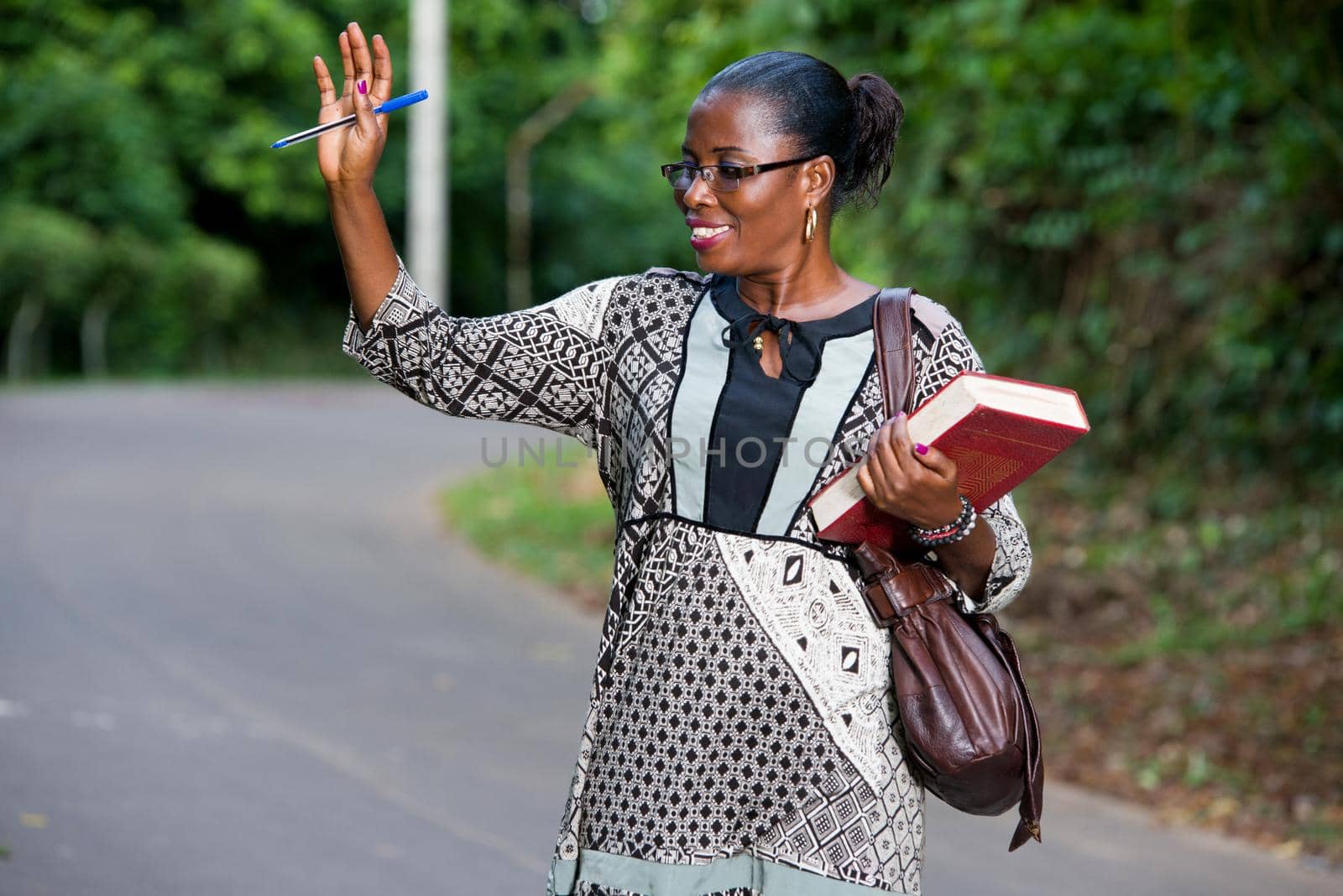 young woman standing in a camisole and holding a book in one hand raises the other hand in greeting with a smile.