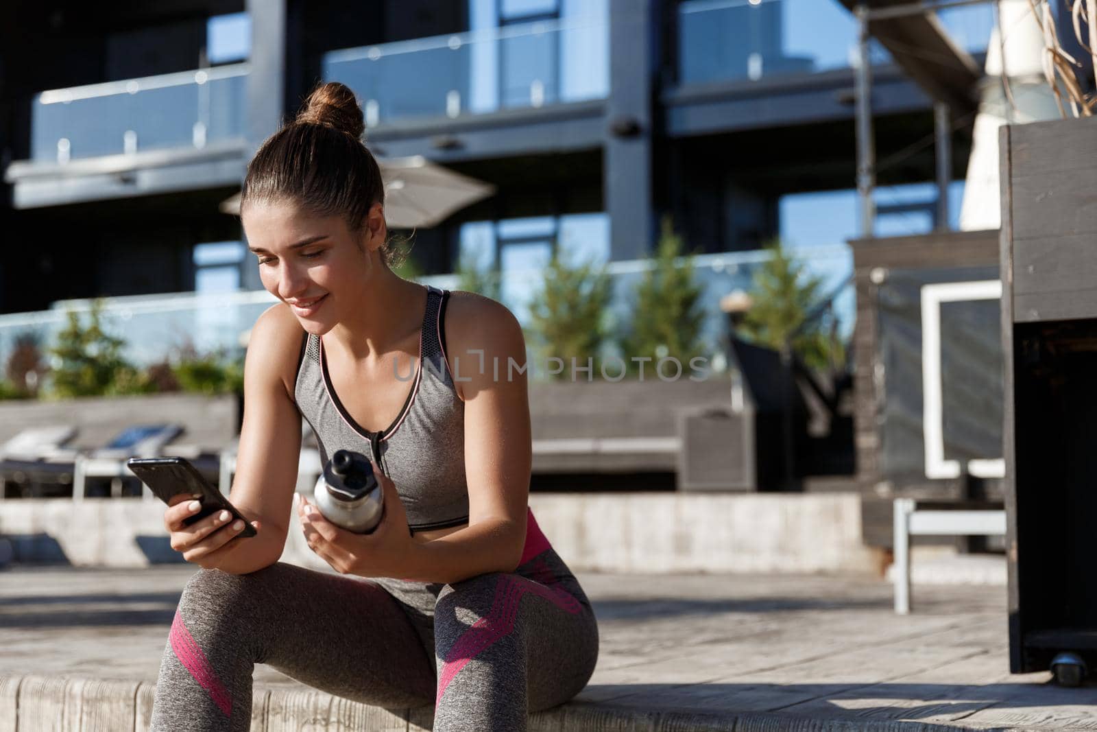 Outdoor shot of attractive sportswoman sitting on bench and using mobile phone, holding water bottle.