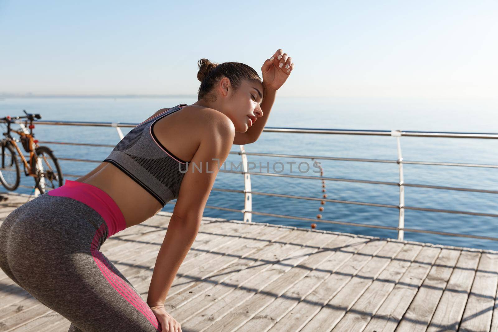 Rear view of attractive fitness woman taking a breath after jogging along seaside, wiping sweat off forehead and panting.