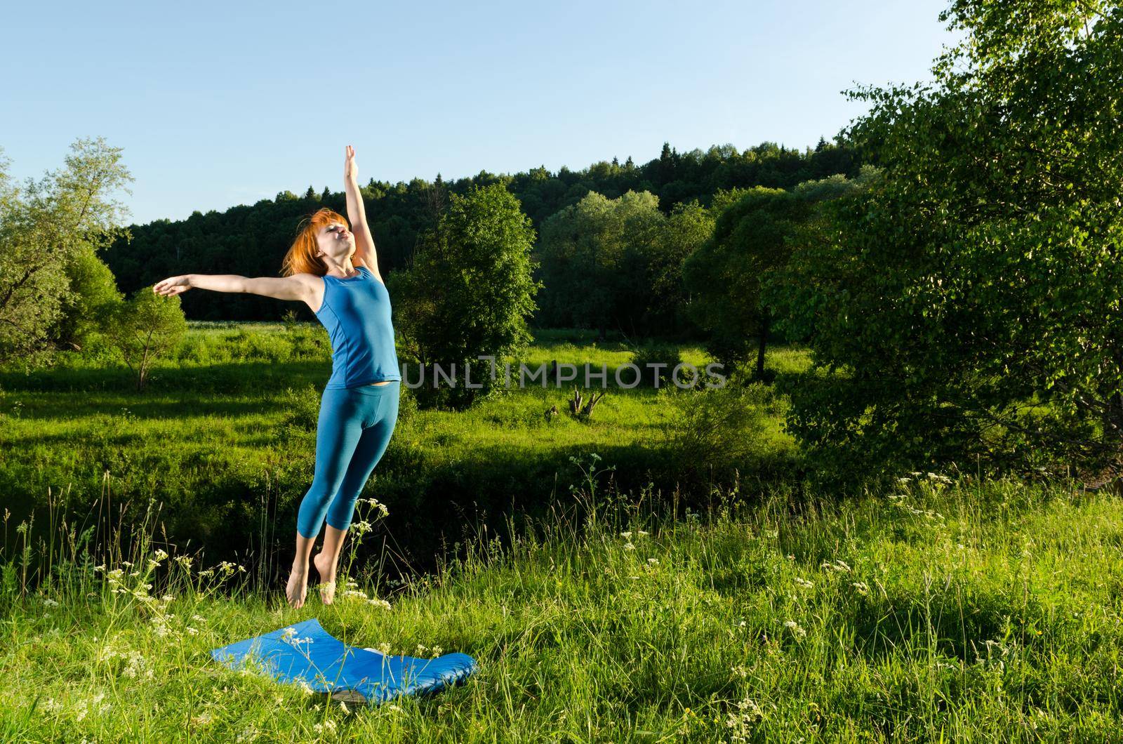 Leaping red haired woman practicing fitness yoga outdoors on a summer day